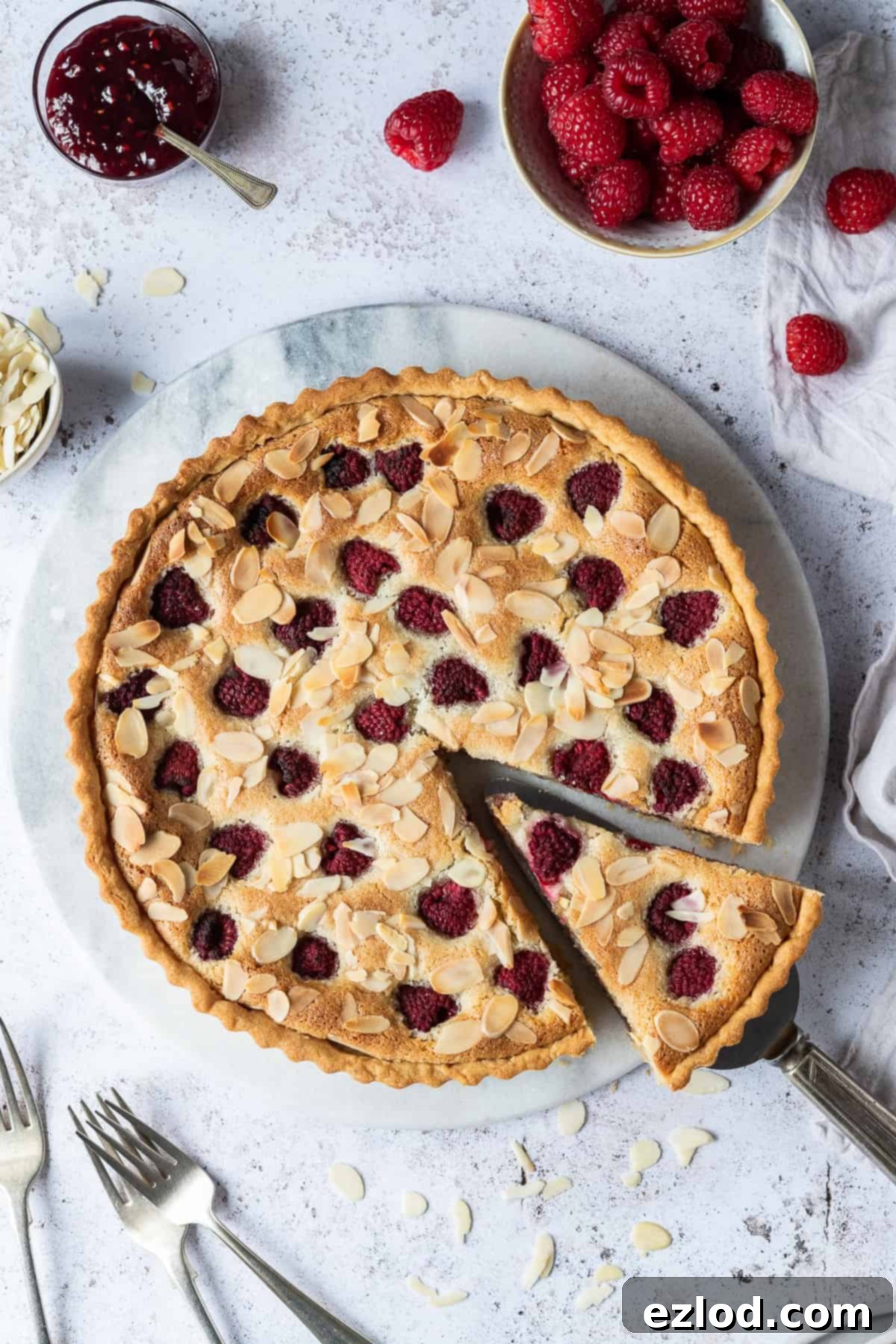 vegan raspberry Bakewell tart on a round marble board on a white background with a bowl of raspberries.