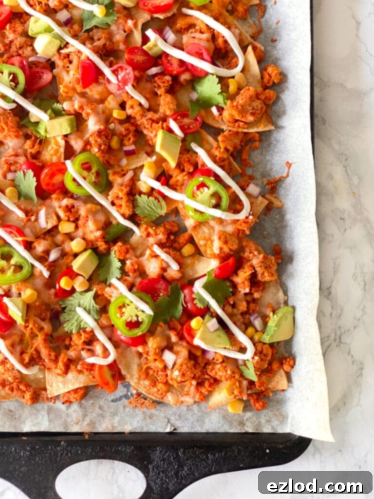 Homemade nachos topped with ground beef, cheese, jalapeno, tomatoes, corn, cilantro, avocado, and drizzled Greek yogurt on a parchment lined sheet pan.