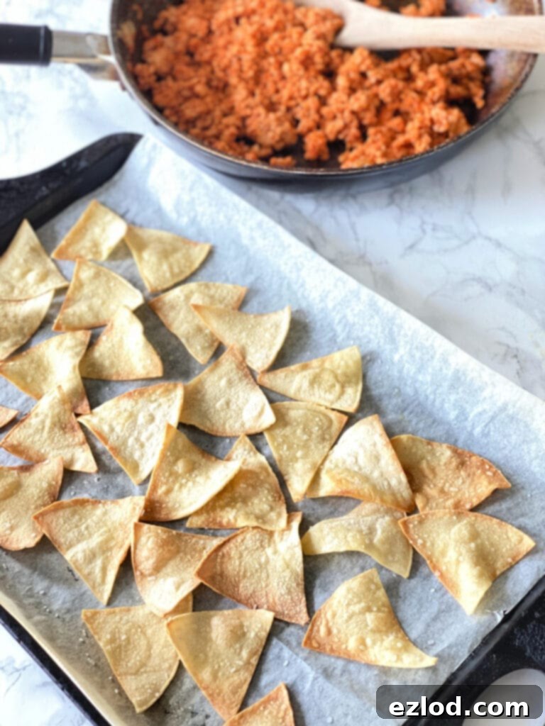 Cooked homemade tortilla chips with browned crispy edges on a parchment lined sheet pan. At the top of the photo is a skillet of cooked ground beef with a wooden spoon,