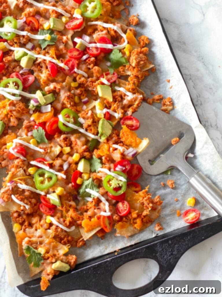 Homemade nachos topped with ground beef, cheese, jalapeno, tomatoes, corn, cilantro, avocado, and drizzled Greek yogurt on a parchment lined sheet pan. In the corner of the sheet pan, some of the nachos have been served and there is a silver utensil resting on the pan.