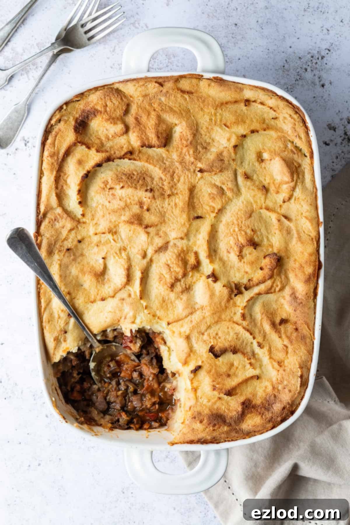 Delicious vegan cottage pie in a white baking dish, presented on a white background with a cream tea towel and two forks, ready to be served.