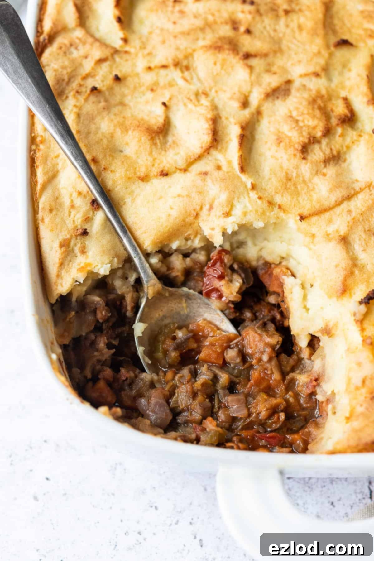 Close-up shot of a steaming vegan cottage pie, with a spoon having just removed a portion, showcasing the rich filling and golden mashed potato topping.