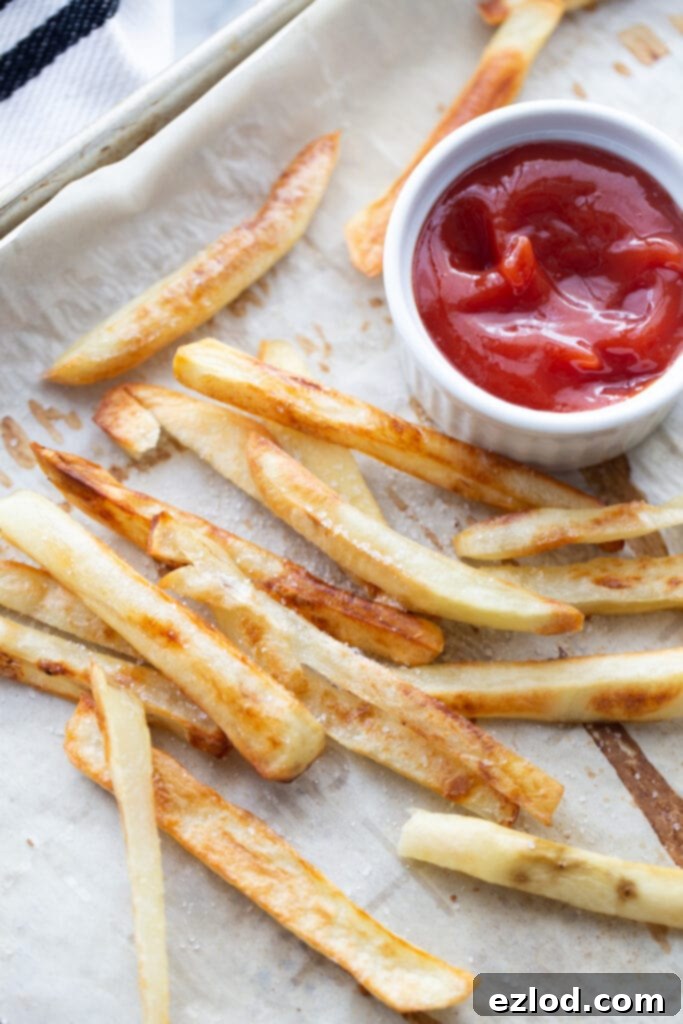 Overhead photo of golden brown homemade baked French Fries on a parchment lined sheet pan. There is a white ramekin of ketchup on the corner of the pan. 