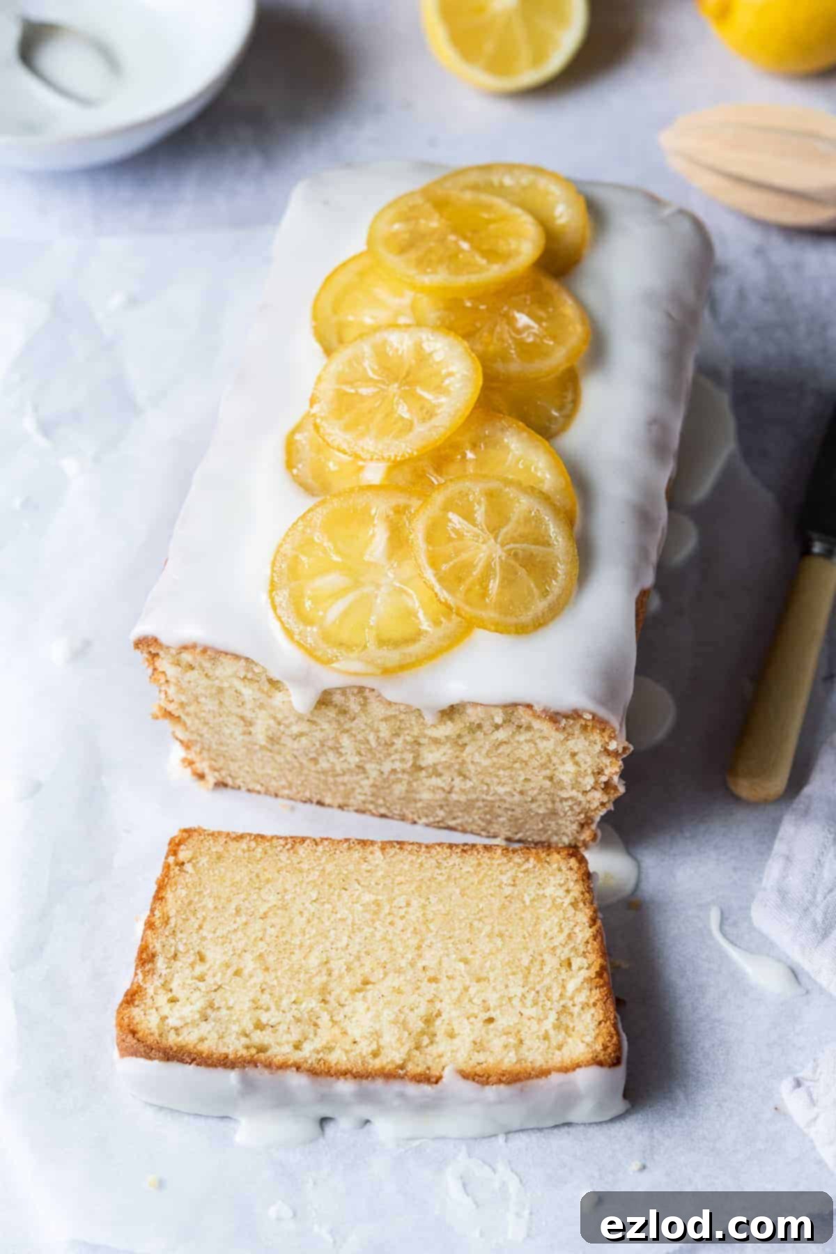 A loaf of vegan lemon pound cake, beautifully topped with delicate candied lemon slices, resting on a sheet of white baking parchment. Fresh lemons and a small bowl of icing are visible in the background.