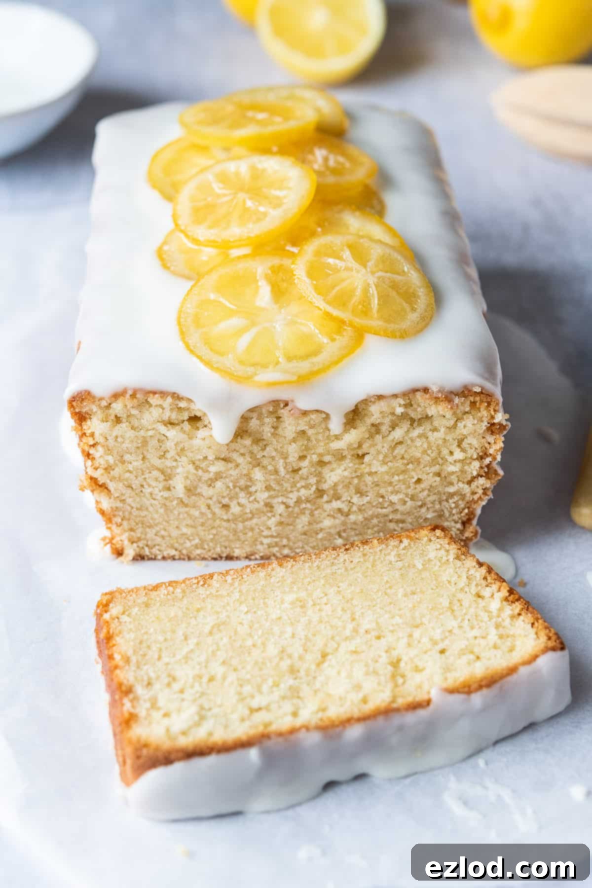 A beautifully presented loaf of vegan lemon pound cake, artfully topped with delicate candied lemon slices. It rests on a clean, white sheet of baking parchment, with fresh lemons and a bowl of creamy icing blurred in the background, creating an inviting culinary scene.