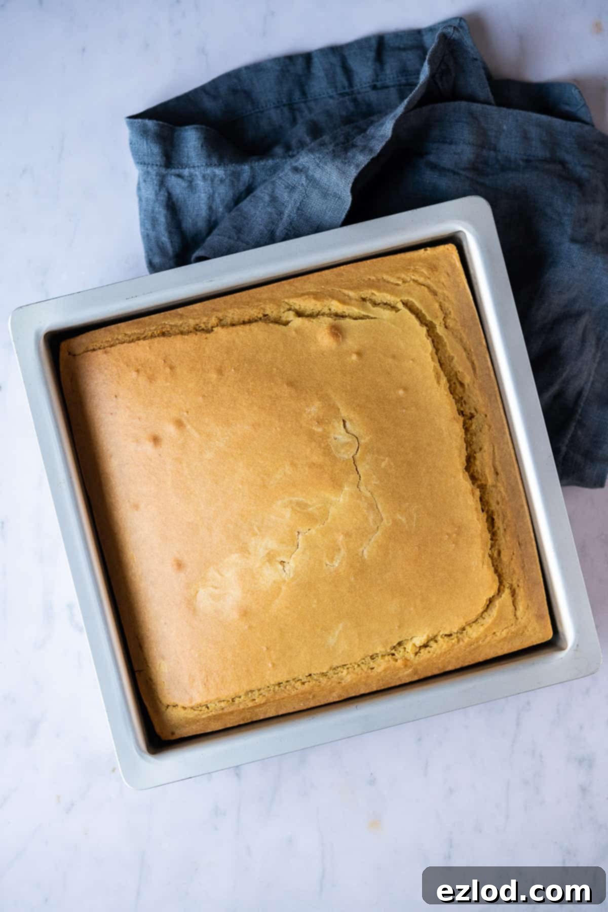 Warm squares of vegan cornbread nestled in a metal cake tin, resting on a elegant marble surface next to a soft grey cloth.