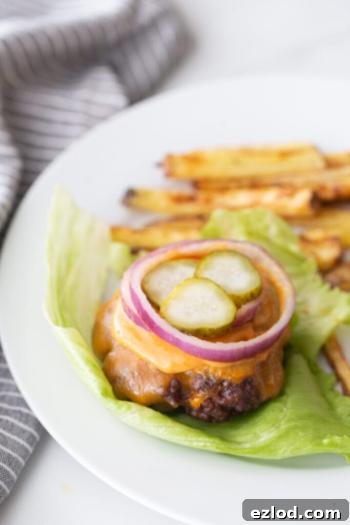 A bunless burger topped with cheddar cheese, burger sauce, red onion, and pickles, on a lettuce leaf. There are french fries on the plate also, out of focus.