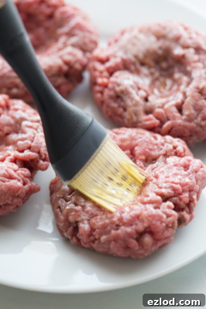 Raw burger patties on a white plate being brushed with olive oil
