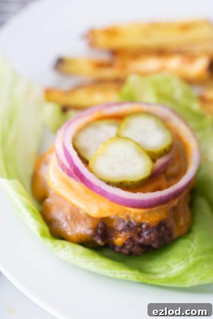 Close of photo of a bunless burger topped with cheddar cheese, burger sauce, red onion, and pickles, on a lettuce leaf. In the background, there are french fries on the plate also, out of focus.