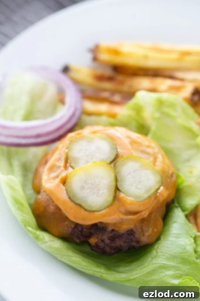 The best bunless burger recipe topped with cheddar cheese, burger sauce,, and pickles, on a lettuce leaf. There are red onions and french fries on the plate also, out of focus.