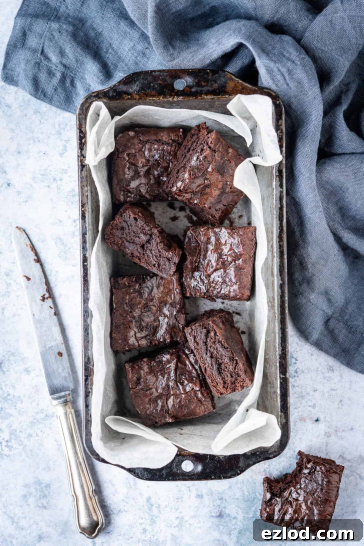 Delicious mini brownie squares nestled in a white baking parchment-lined loaf tin on a sleek grey background, accompanied by a soft grey cloth. One brownie, with a tempting bite taken out, highlights its fudgy interior.
