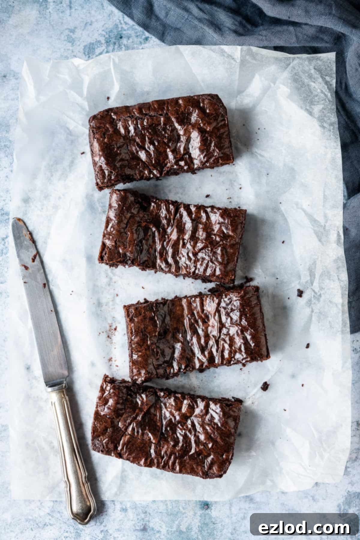 Four perfectly portioned small batch vegan brownies arranged on a sheet of white baking parchment, resting on a sophisticated grey background. A knife and a soft grey cloth are subtly placed beside them.