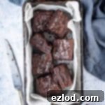 Mini brownie squares in a white baking parchment lined loaf tin on a grey background with a grey cloth, one brownie with a bite taken out of it.