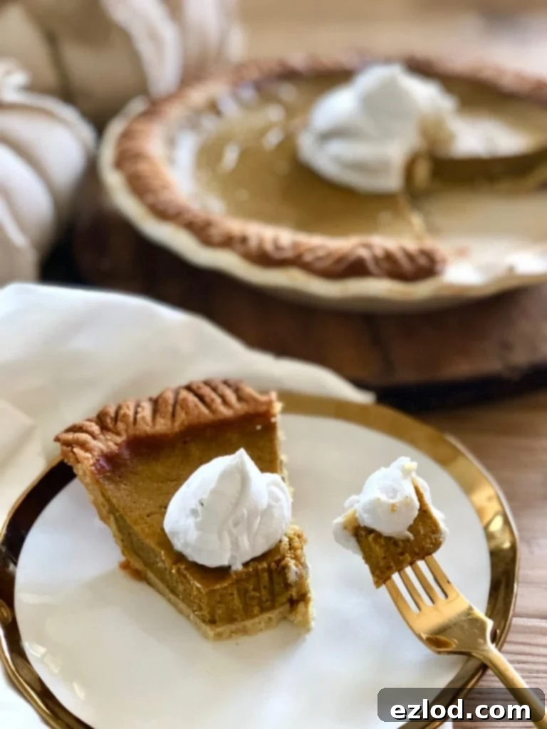 A slice of Healthy Pumpkin Pie, topped with light coconut cream, rests on a white plate with a gold rim. A gold fork holds a bite-sized piece of pie, with the full pie blurred in the background, adorned with coconut cream.