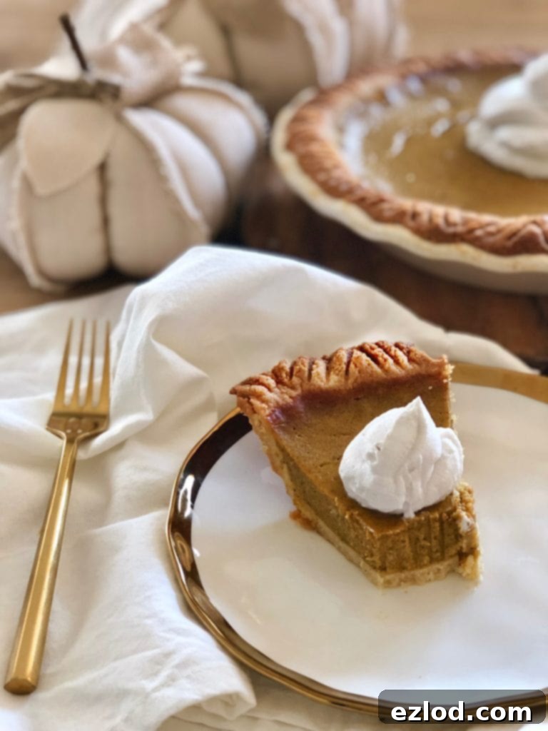 A slice of Healthy Pumpkin Pie with coconut cream sits on a white, gold-rimmed plate. Beside it, a gold fork rests on a white fabric napkin. The rest of the pie is visible in the soft focus background.