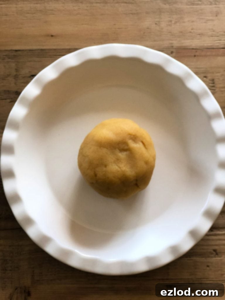 A ball of almond flour pie crust dough is centered in a white pie plate, resting on a rustic wooden background, ready to be pressed.