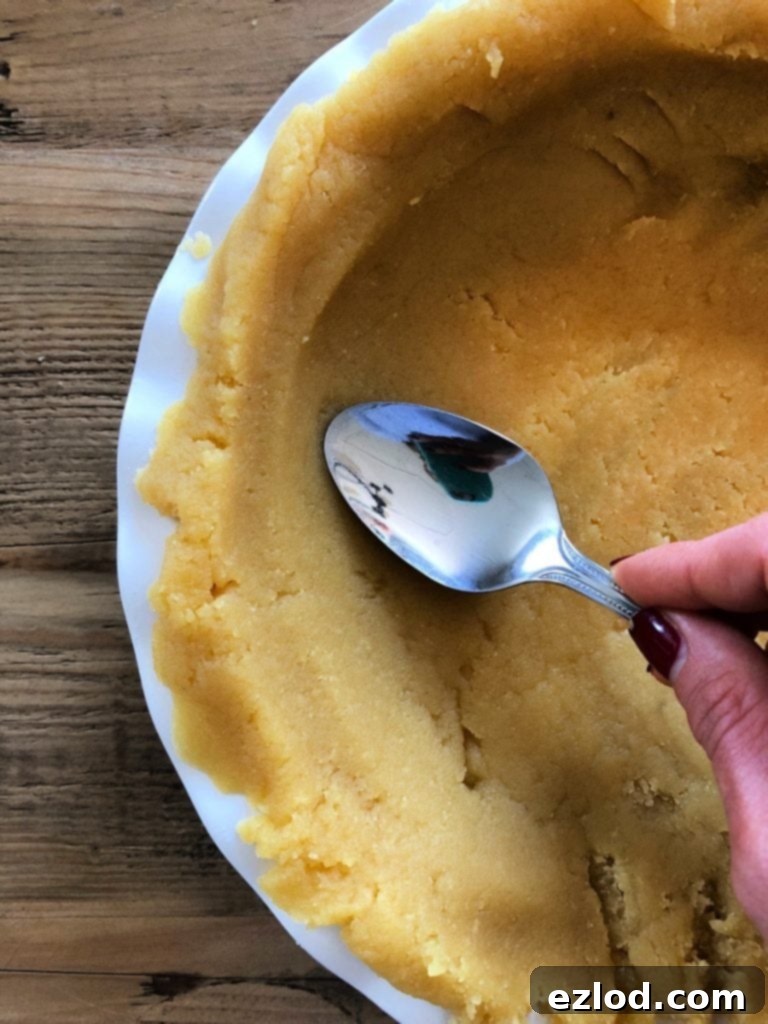 A spoon is gently pressing down on the raw almond flour crust that lines a white pie plate, preparing it for baking. The plate is on a wooden background.