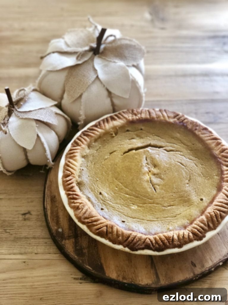A beautifully baked Healthy Pumpkin Pie sits in a white pie plate on a wooden pedestal, surrounded by two cream-colored fabric pumpkins on a wooden table, evoking a cozy autumn scene.