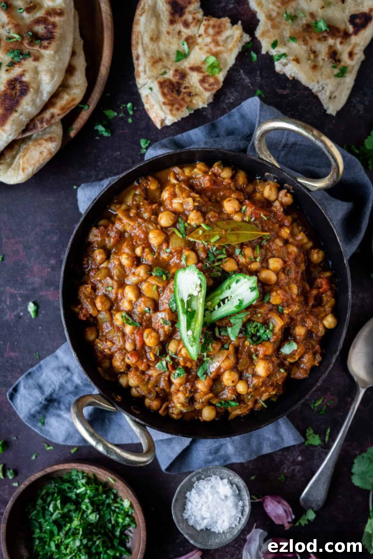 Chana masala in a black metal dish on a dark background with naan bread, fresh coriander, a pot of salt and a spoon.