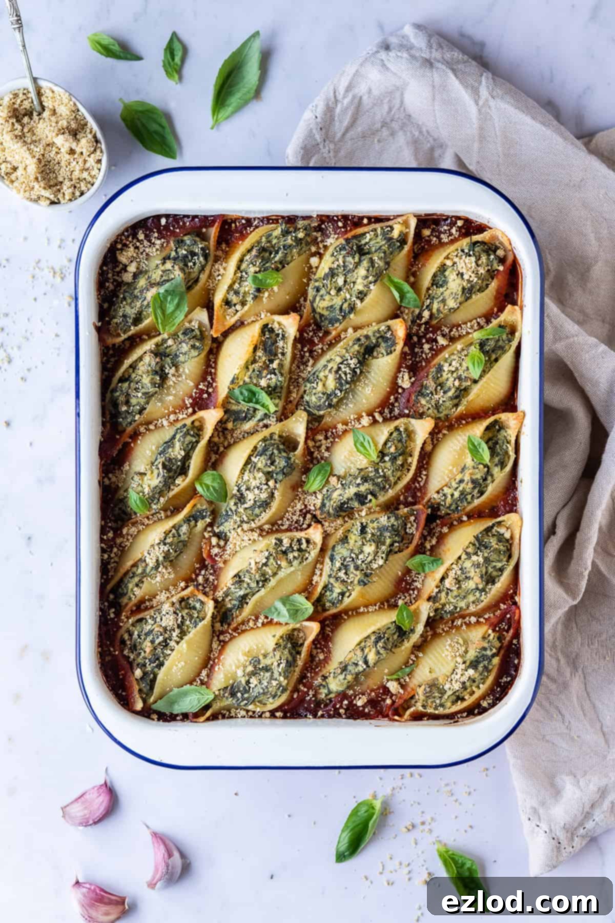 Delicious vegan spinach and almond tofu ricotta stuffed pasta shells garnished with fresh basil and vegan parmesan, served in a white enamel baking dish on a rustic marble background.