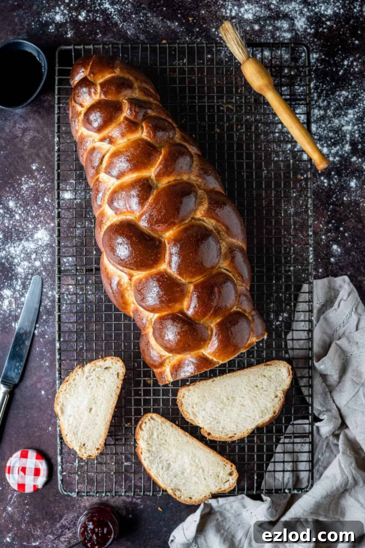A beautifully sliced loaf of vegan challah bread resting on a wire cooling rack, set against a dark rustic background. Nearby, a cloth, a pot of homemade jam, and a pastry brush add to the cozy baking scene. The bread shows its soft, airy crumb.