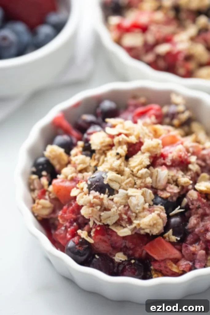 A vibrant mixed berry crisp with a gluten-free oat topping, served in a shallow white ramekin. In the soft-focus background, two more ramekins hold blueberry crisp, garnished with fresh strawberries and raspberries, hinting at the delicious variety.