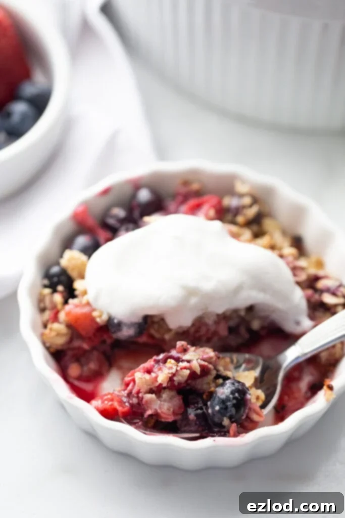 An individual serving of mixed berry crisp, generously topped with a dollop of homemade coconut cream. A spoon with a tantalizing bite of crisp waits, ready to be enjoyed.