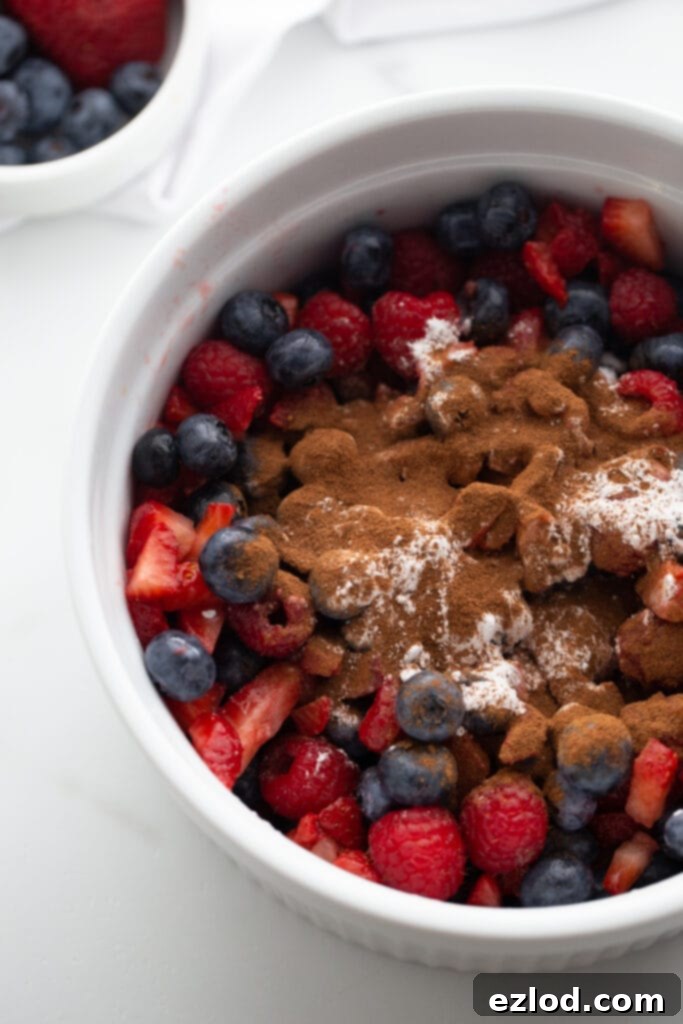 A close-up view of a large white casserole dish brimming with fresh blueberries, sliced strawberries, and vibrant raspberries. The berries are gently dusted with cinnamon and arrowroot powder, perfectly prepped and ready for the Instant Pot to transform them into a delicious crisp.