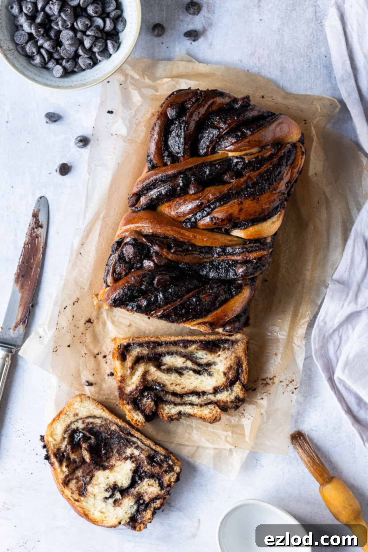 A beautifully baked vegan chocolate babka loaf, accompanied by a bowl of chocolate chips, a knife, and a pastry brush, all resting on brown baking parchment against a grey background.