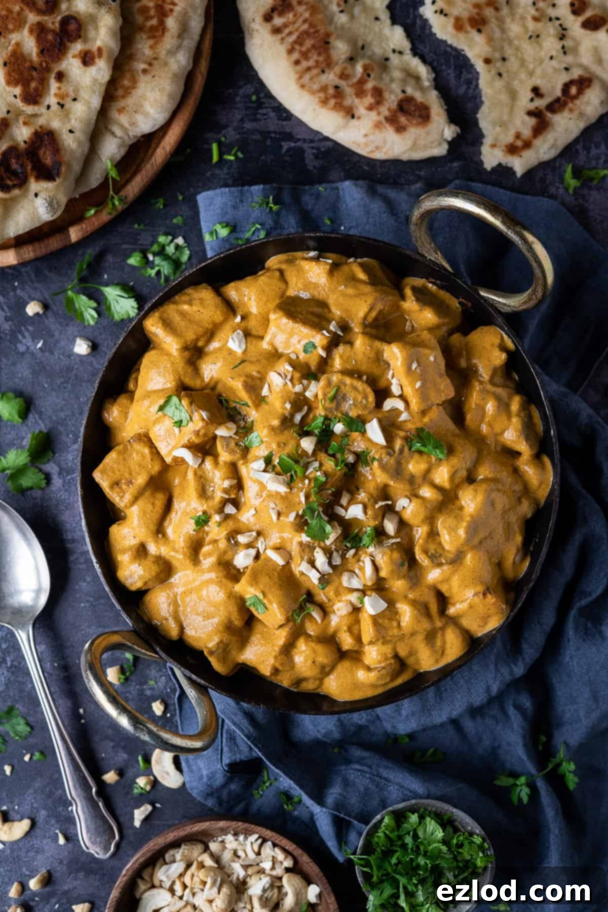Vegan mushroom and tofu korma in a black karahi pan on a dark background with chopped cashews, coriander and naan breads.