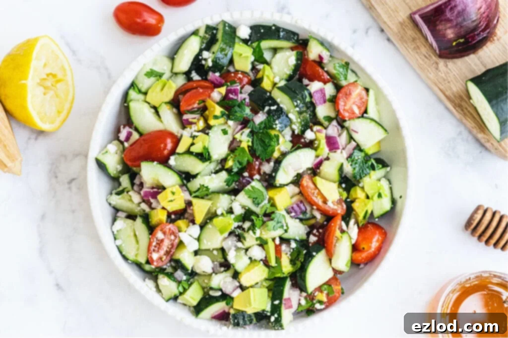 A large white bowl of summer cucumber tomato salad topped with avocado and feta cheese with other ingredients on the side of the bowl.