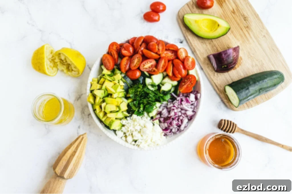Overhead photo of large bowl of cucumber tomato salad before being mixed together. Extra ingredients surround the bowl of salad.