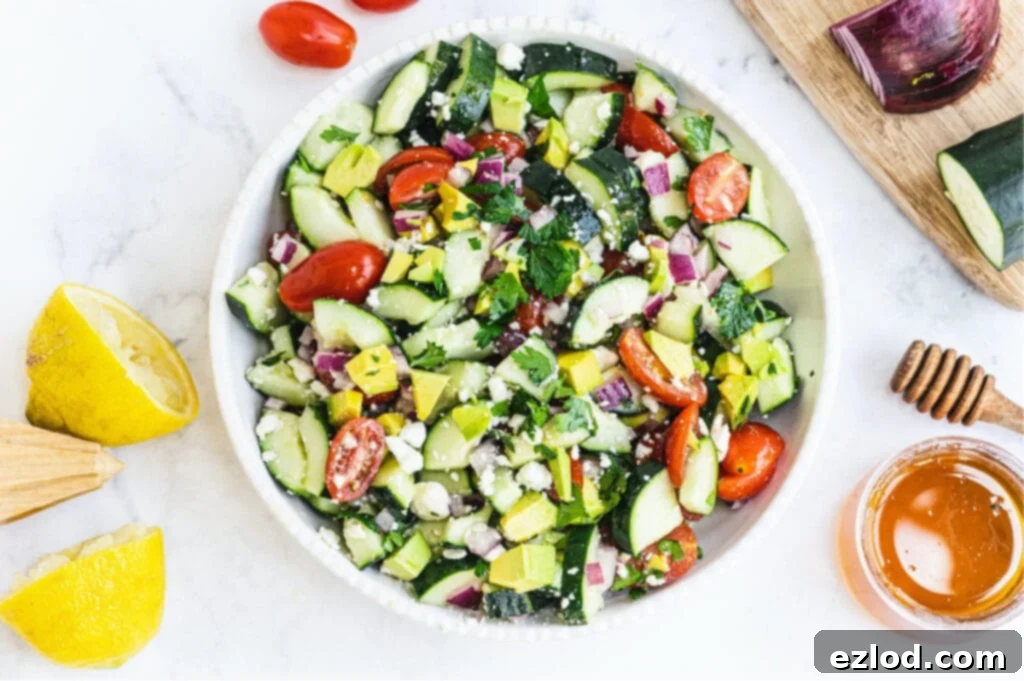 Salad made of tomatoes, cucumbers, red onion, feta, and avocado in a large white serving bowl. Lemons, honey, tomatoes, red onion, and cucumber all surround the photo at the edges.