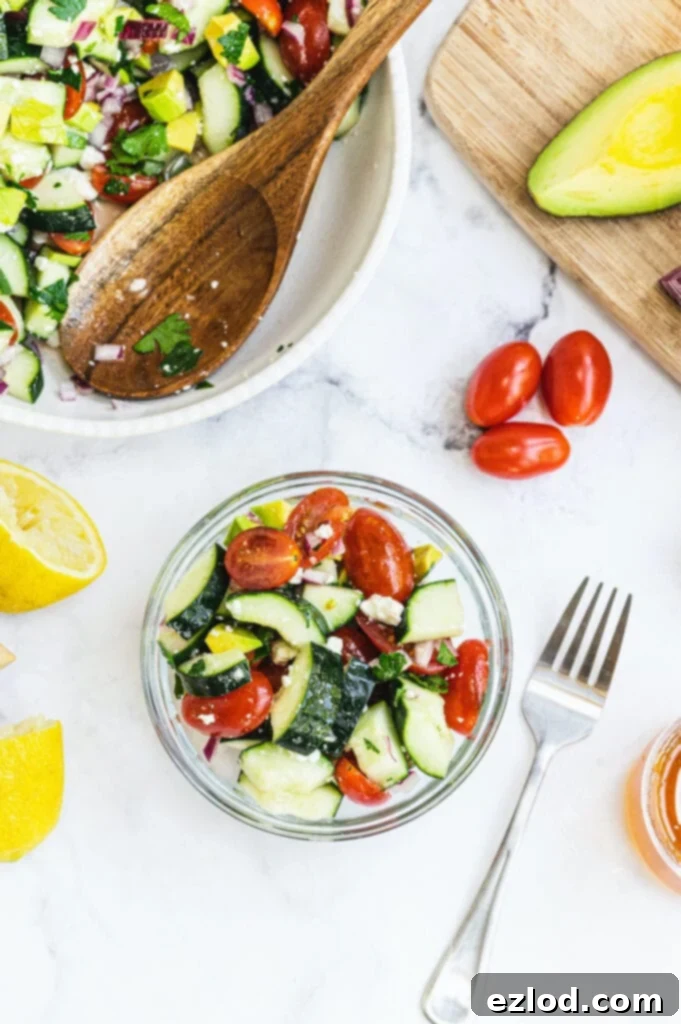 Cucumber tomato feta salad in a small glass bowl with a large serving bowl full of salad off to the side. 