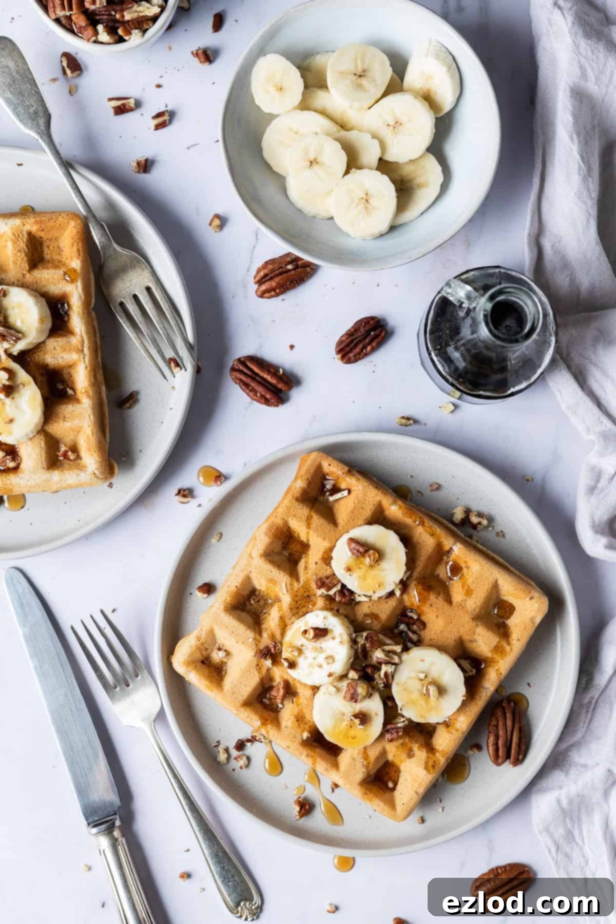 two plates of vegan banana waffles, each topped with sliced banana, pecans, and maple syrup, set on a elegant marble background.