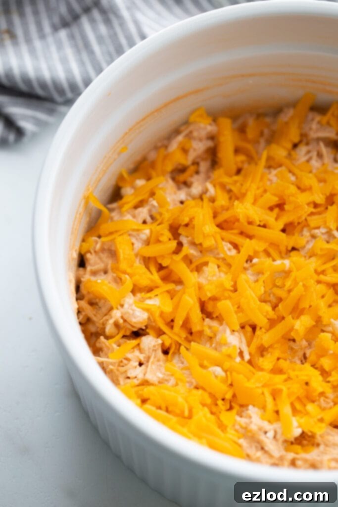 White casserole dish with buffalo chicken dip topped with shredded cheddar cheese, ready for the oven to become golden and bubbly.