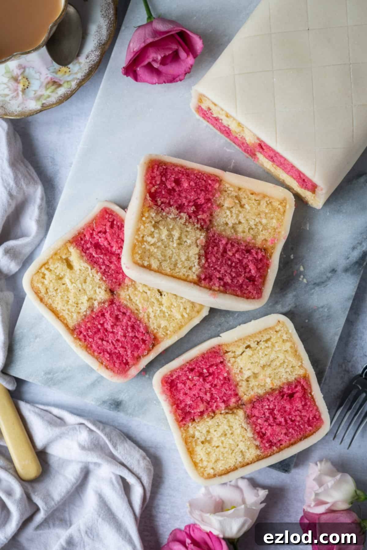 Three perfectly sliced pieces of vegan Battenberg cake on a pristine marble serving board, adorned with delicate fresh roses and a steaming cup of tea in the background. The cake showcases its distinct pink and yellow chequered pattern, encased in smooth marzipan, highlighting its tender crumb and inviting texture.