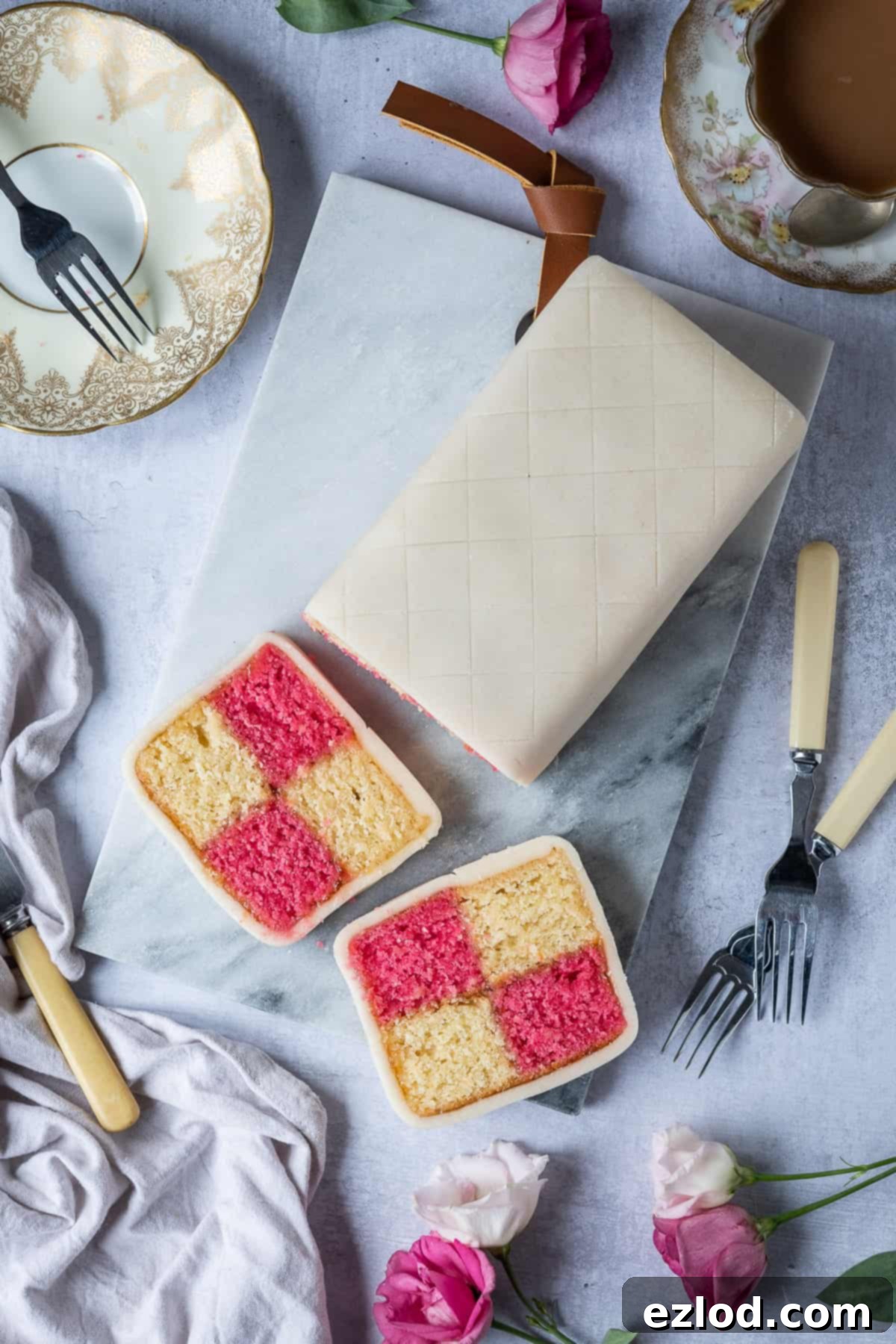 A beautifully styled scene featuring several slices of vegan Battenberg cake on a marble board, surrounded by fresh flowers, a grey cloth napkin, delicate pastry forks, and a steaming cup of tea, evoking an elegant afternoon tea setting.