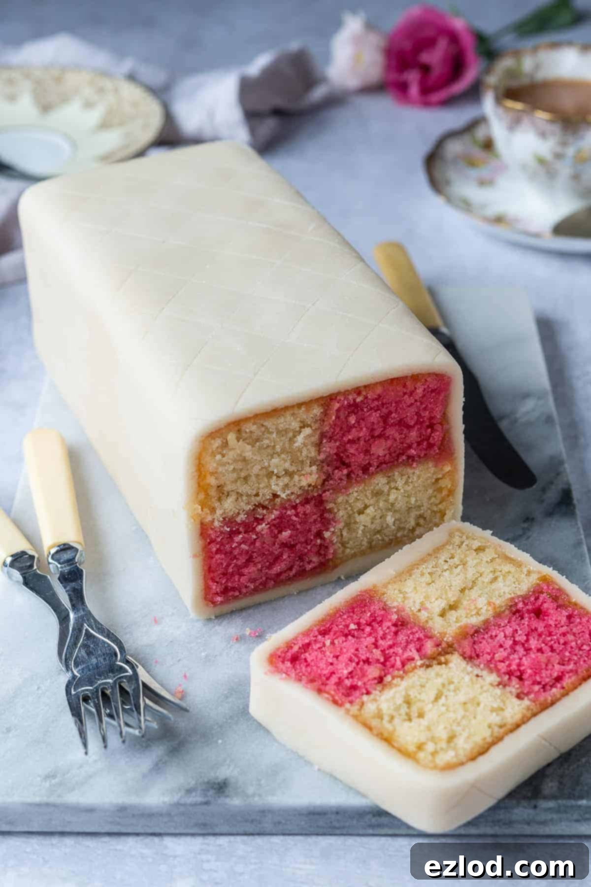 A beautifully presented vegan Battenberg cake resting on a sleek marble board, set against a subtle grey background. The cake is whole, showcasing the outer marzipan layer, ready to be sliced and reveal its colourful interior.