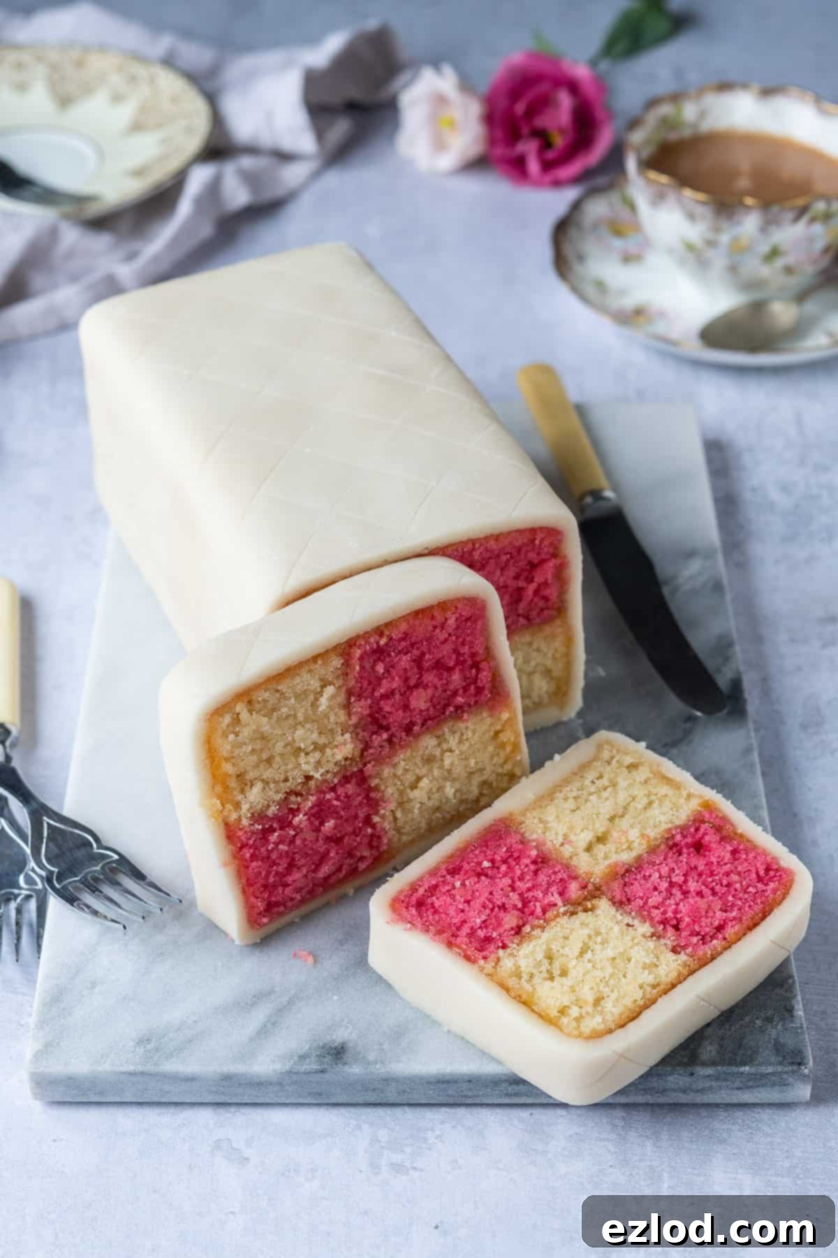 A whole vegan Battenberg cake, perfectly wrapped in marzipan, presented on a marble board. The ends have been neatly trimmed to showcase the striking pink and yellow chequered pattern.