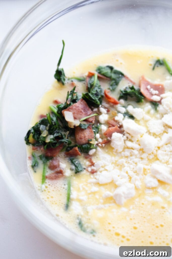Uncooked eggs, fresh spinach, diced bacon, and crumbled feta cheese are mixed in a clear glass bowl, ready for the Instant Pot frittata recipe.
