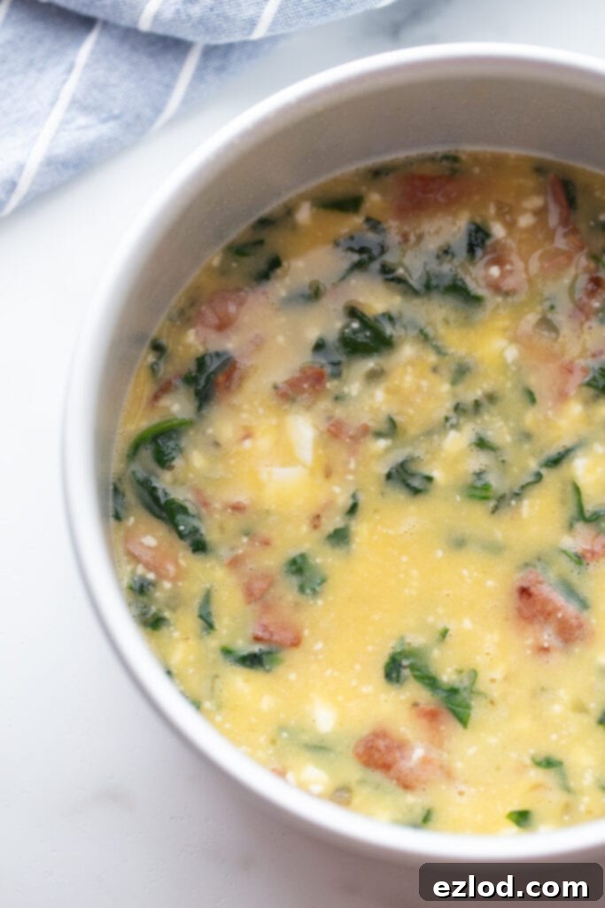 An overhead view of scrambled egg mixture with vibrant spinach, crispy bacon, and creamy feta cheese, perfectly portioned in a round baking pan, ready for cooking.
