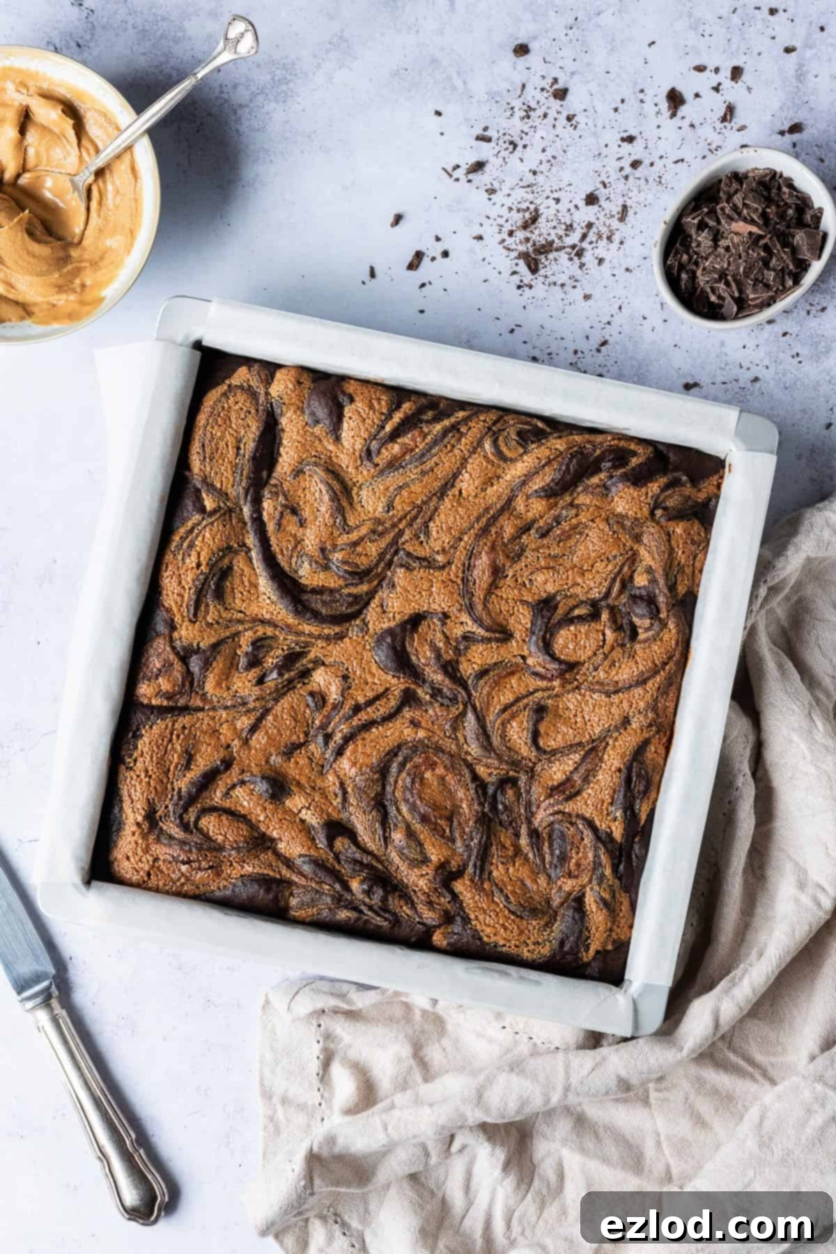 A pan of vegan peanut butter swirl brownies on a grey background with a cloth, bowl of peanut butter and bowl of chopped chocolate.