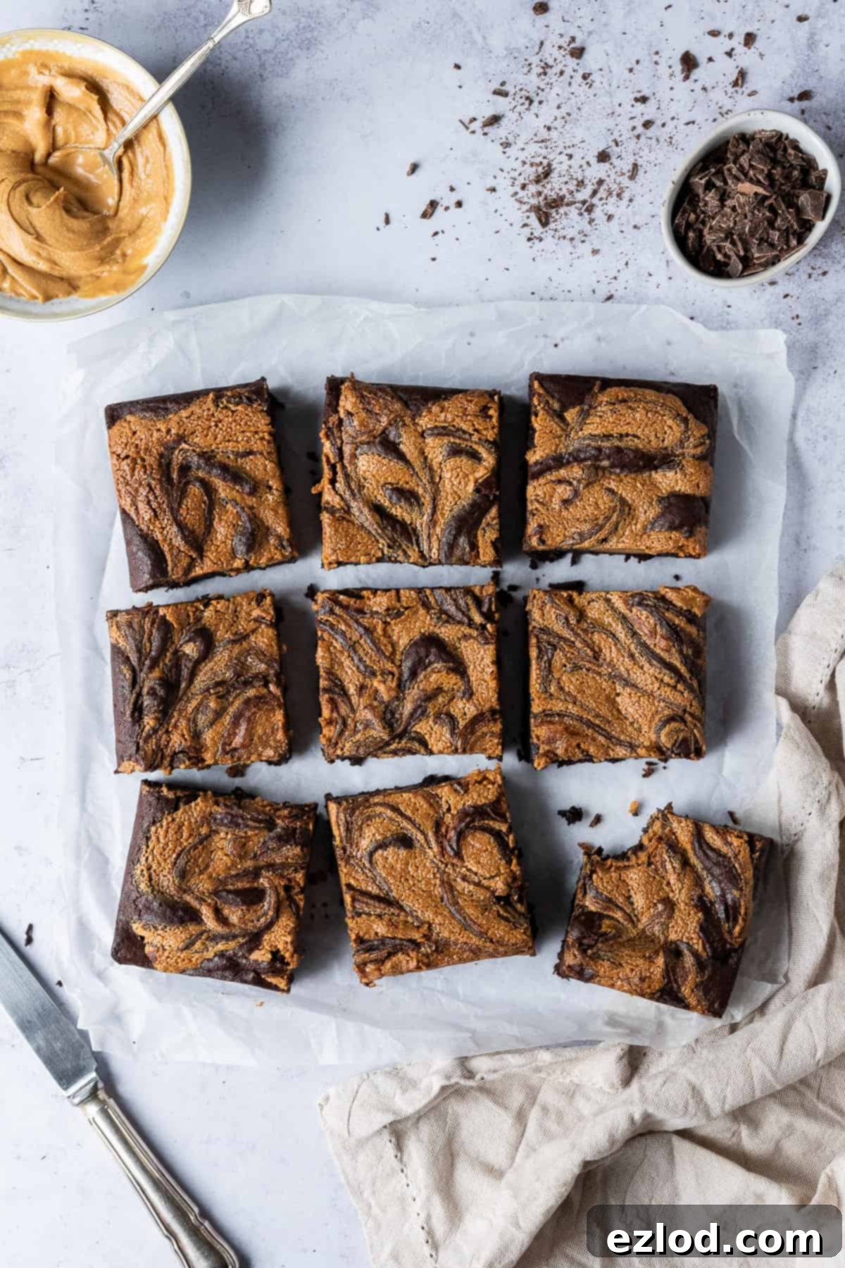 Squares of vegan peanut butter swirl brownie on a sheet of baking parchment with a cloth, a bowl of peanut butter and a bowl of chopped chocolate.