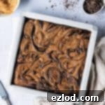 A pan of vegan peanut butter swirl brownies on a grey background with a cloth, bowl of peanut butter and bowl of chopped chocolate.