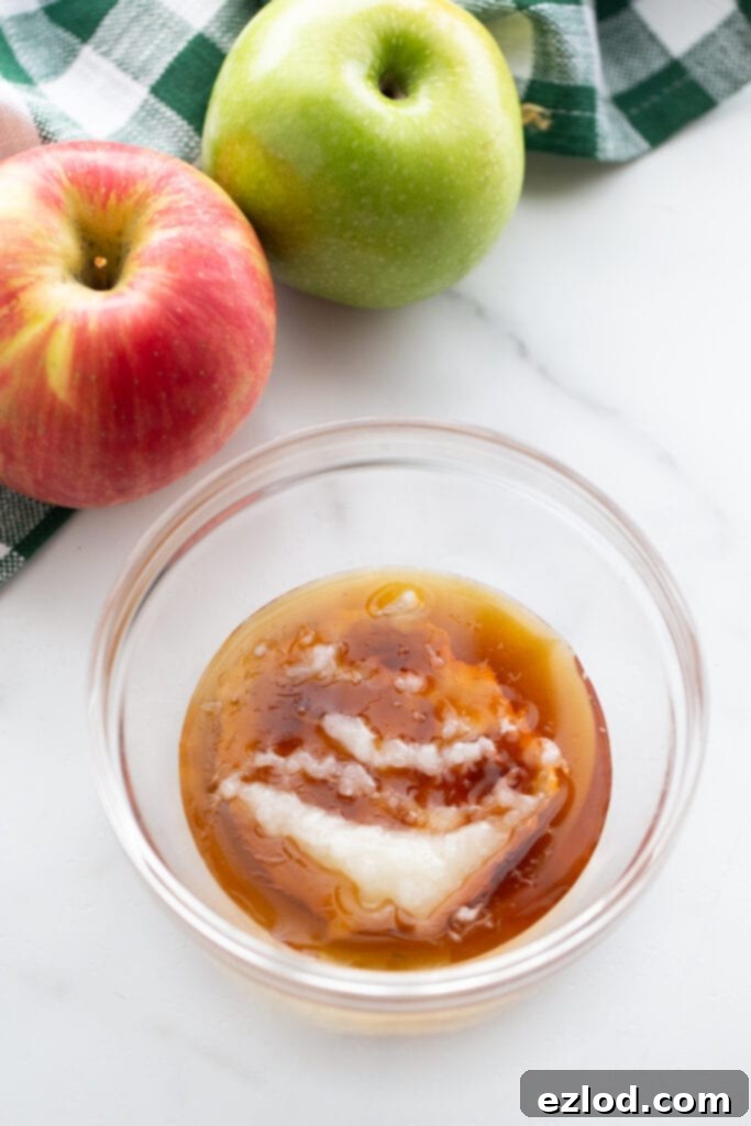 Small glass bowl containing melted maple syrup and coconut oil, ready to be microwaved for caramel dip. Two whole red apples are placed beside the bowl.