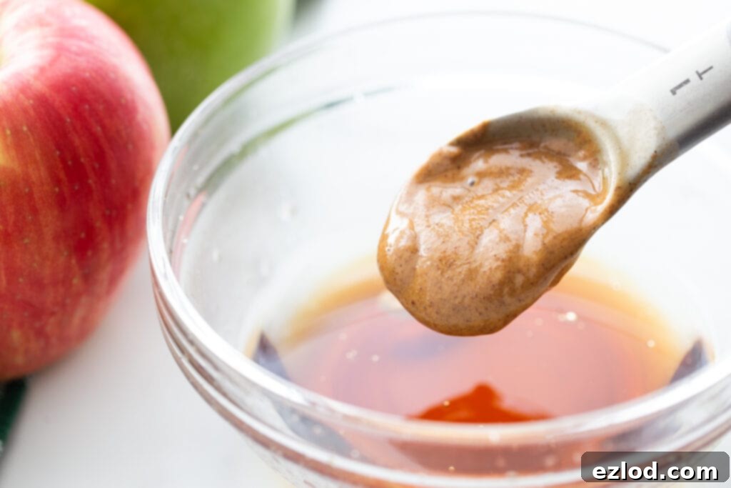 A measuring spoon full of creamy almond butter being carefully added to a glass bowl of brown liquid, part of the caramel making process.