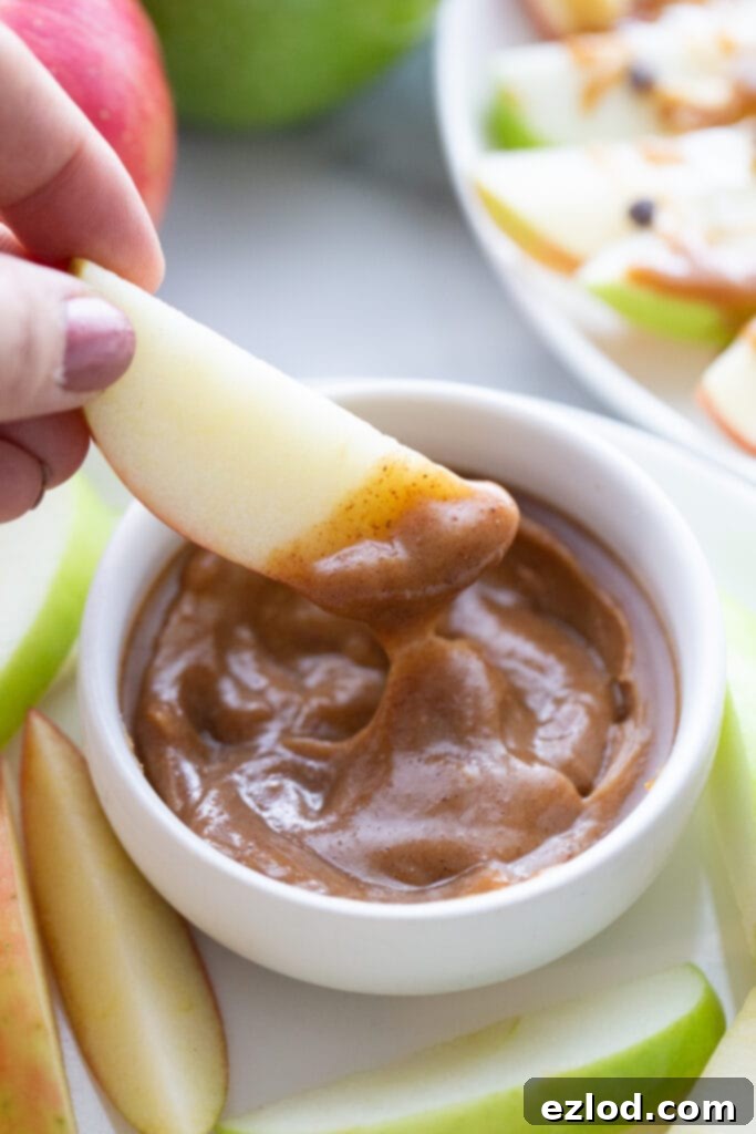 A hand holding a slice of crisp apple dipped generously in rich, homemade vegan caramel. The caramel bowl is surrounded by more apple slices, with a plate of apple nachos blurred in the background.