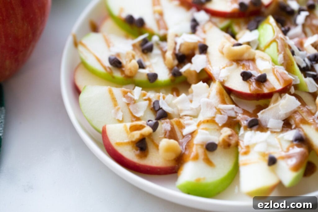 Close-up of a plate of vibrant apple nachos, featuring sliced red and green apples generously topped with shredded coconut, mini chocolate chips, chopped walnuts, and a luscious drizzle of homemade caramel sauce.