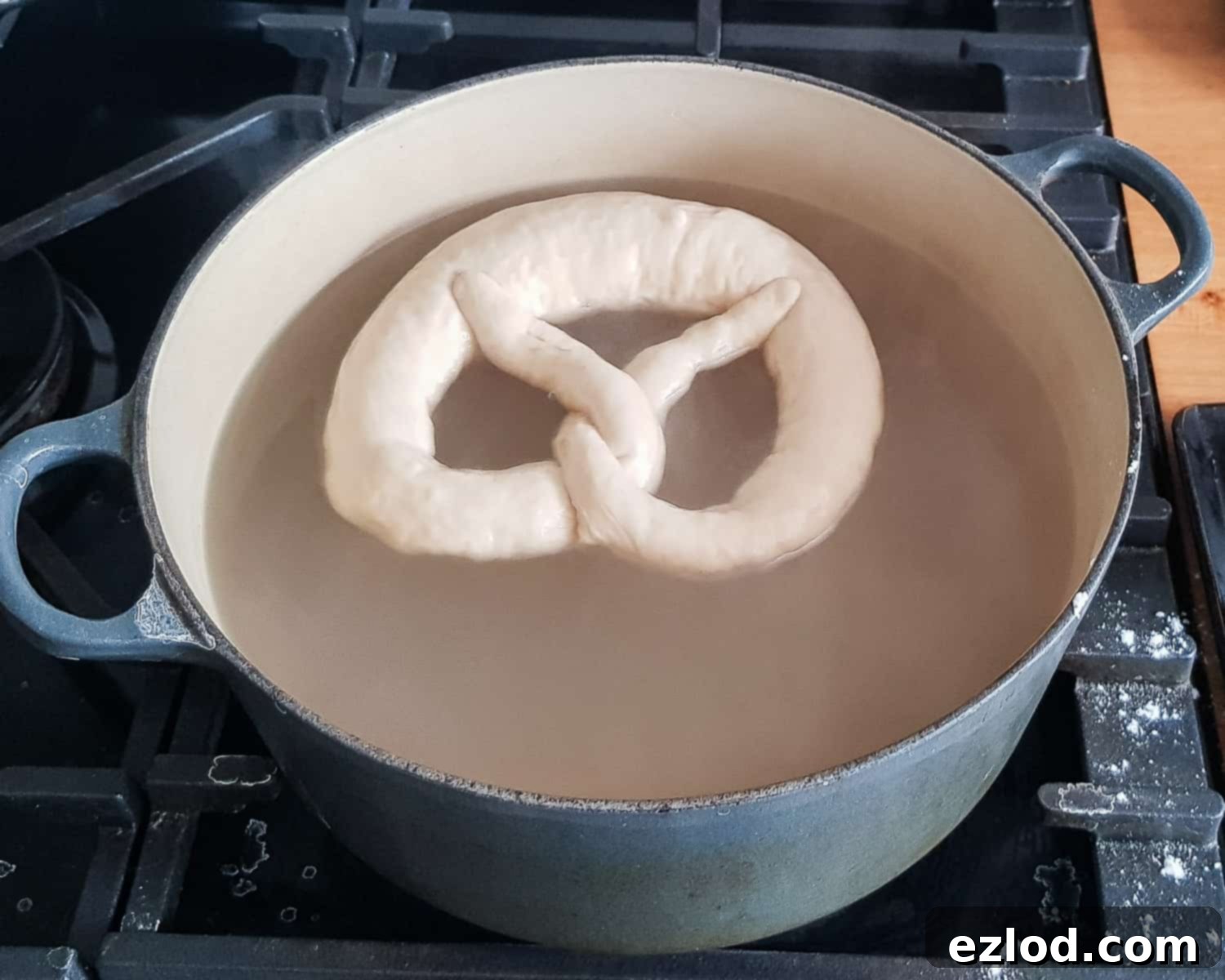 Step-by-step image showing a pretzel being carefully poached in a pan of bicarbonate of soda solution, with a ladle pouring the solution over it.